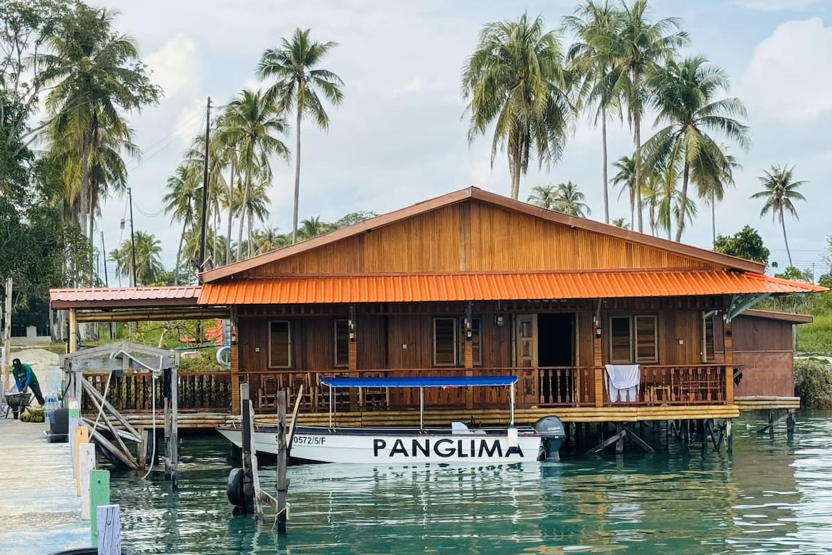 Una casa flotante de madera con techo rojo, amarrada en aguas tranquilas, rodeada de palmeras.