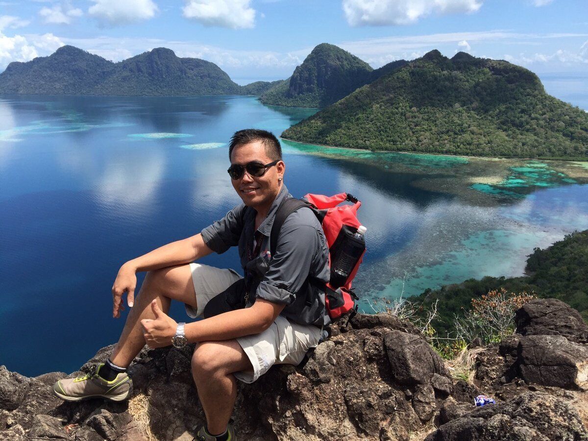 A person sitting on a rocky outcrop overlooking a large body of water with lush greenery and mountains in the background.