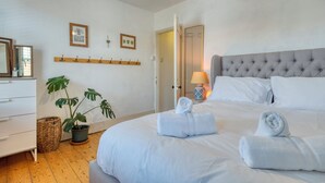 A neatly arranged bedroom with a large bed, a bedside table with a lamp, a potted plant, and a wooden shelf with hooks.