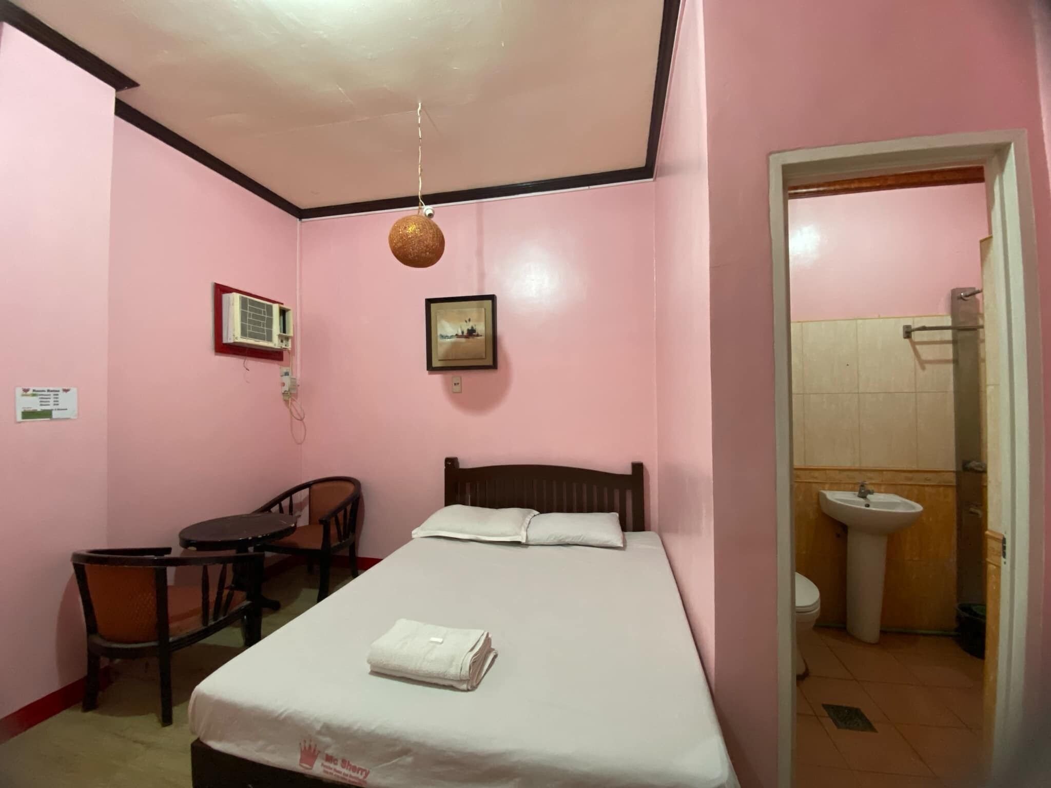 Una habitación rosa con una cama, un escritorio, una silla, un cuadro en la pared y un baño visible a través de una puerta abierta.