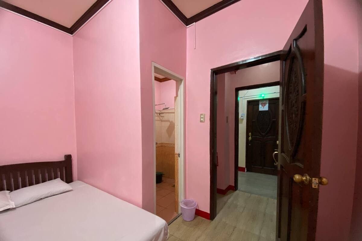 A room with pink walls, a wooden bed, a door leading to another room, and a visible trash bin.