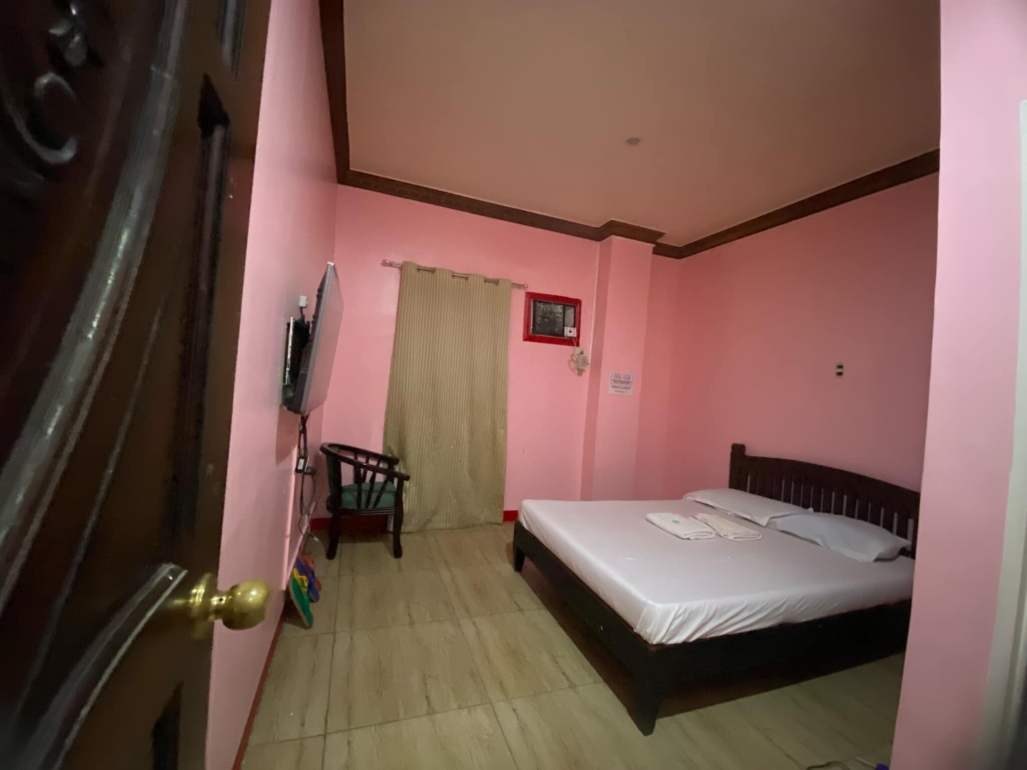 A room with pink walls, a bed with white linens, a wooden chair, a curtain, and a television on a stand.