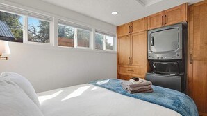 A bedroom with a bed, a blue blanket, a set of towels, wooden cabinets, a built-in oven, and a window with a view of trees. - Charming Historical Home in Downtown Boise (Boise)