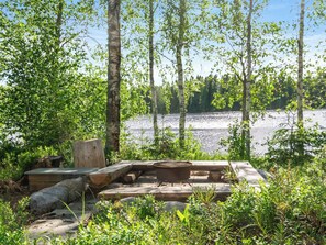 Un espace extérieur doté d’un foyer, de bancs en bois et offrant une vue sur un lac entouré d’arbres.