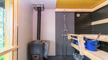 A sauna room with a wood-burning stove, a shower, and a wooden bench with a bucket.