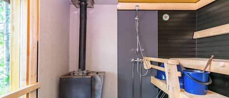 A sauna room with a wood-burning stove, a shower, and a wooden bench with a bucket.