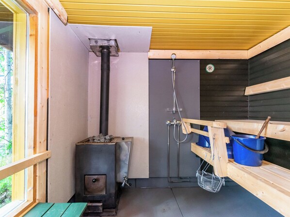 A sauna room with a wood-burning stove, a shower, and a wooden bench with a bucket.