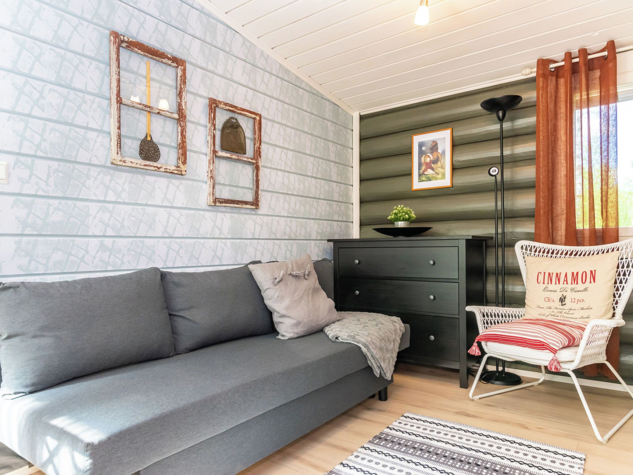 A cozy room with a grey sofa, a small black dresser, a white wicker chair with a cushion, and a patterned rug.
