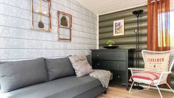 A cozy room with a grey sofa, a small black dresser, a white wicker chair with a cushion, and a patterned rug.