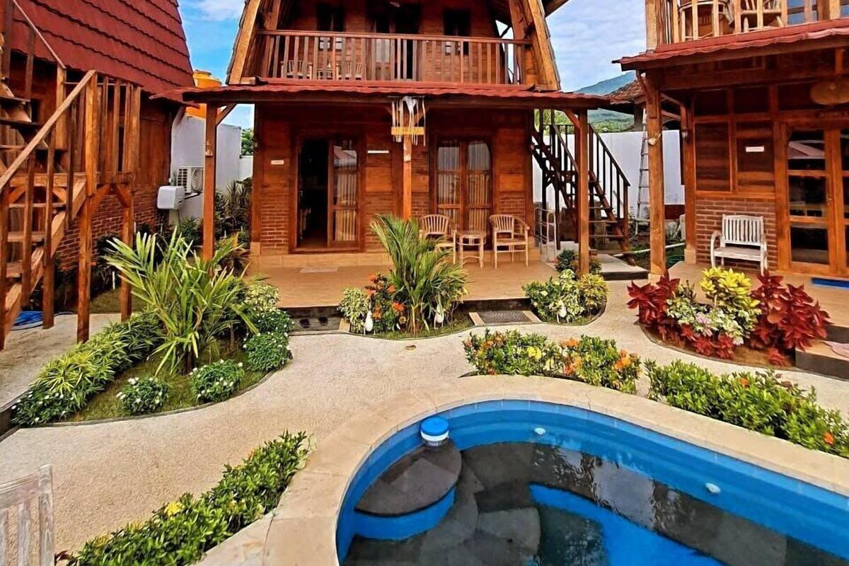 A wooden cabin with a balcony, a pool, and a patio area with plants.