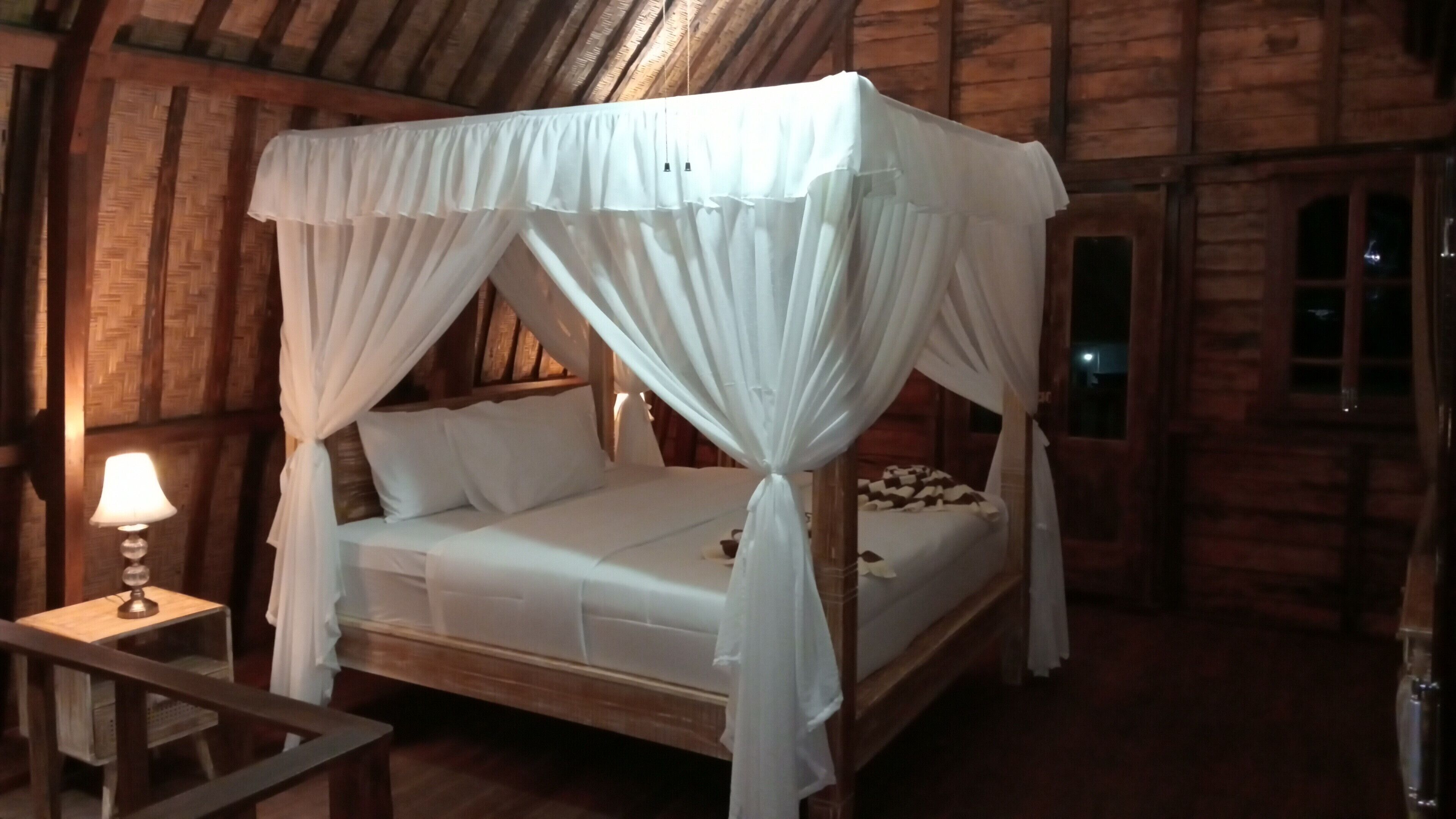 A wooden cabin bedroom with a four-poster bed, white bedding, and a nightstand with a lamp.