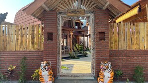 Ein rustikales Holzgebäude mit einem 'WELCOME'-Schild am Eingang, zwei Tigerstatuen und Blick ins Innere.