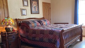 A bedroom with a wooden bed, a nightstand with a lamp, a patterned rug, and a ceiling fan. - Quaint, handicap accessible house in serene Lehigh, classified as a ghost town. (Lehigh)