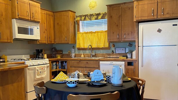 A kitchen with wooden cabinets, a microwave, a stove, and a dining table set for two. - Quaint, handicap accessible house in serene Lehigh, classified as a ghost town. (Lehigh)