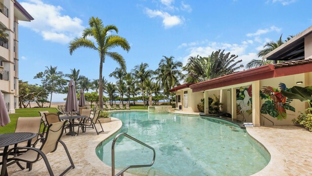 A swimming pool with lounge chairs and umbrellas, surrounded by palm trees and a building with a red roof. - Jaco Beachfront Elegance at Tres Regalos Pristine Views in Lovely Jacó Beach (Jacó)