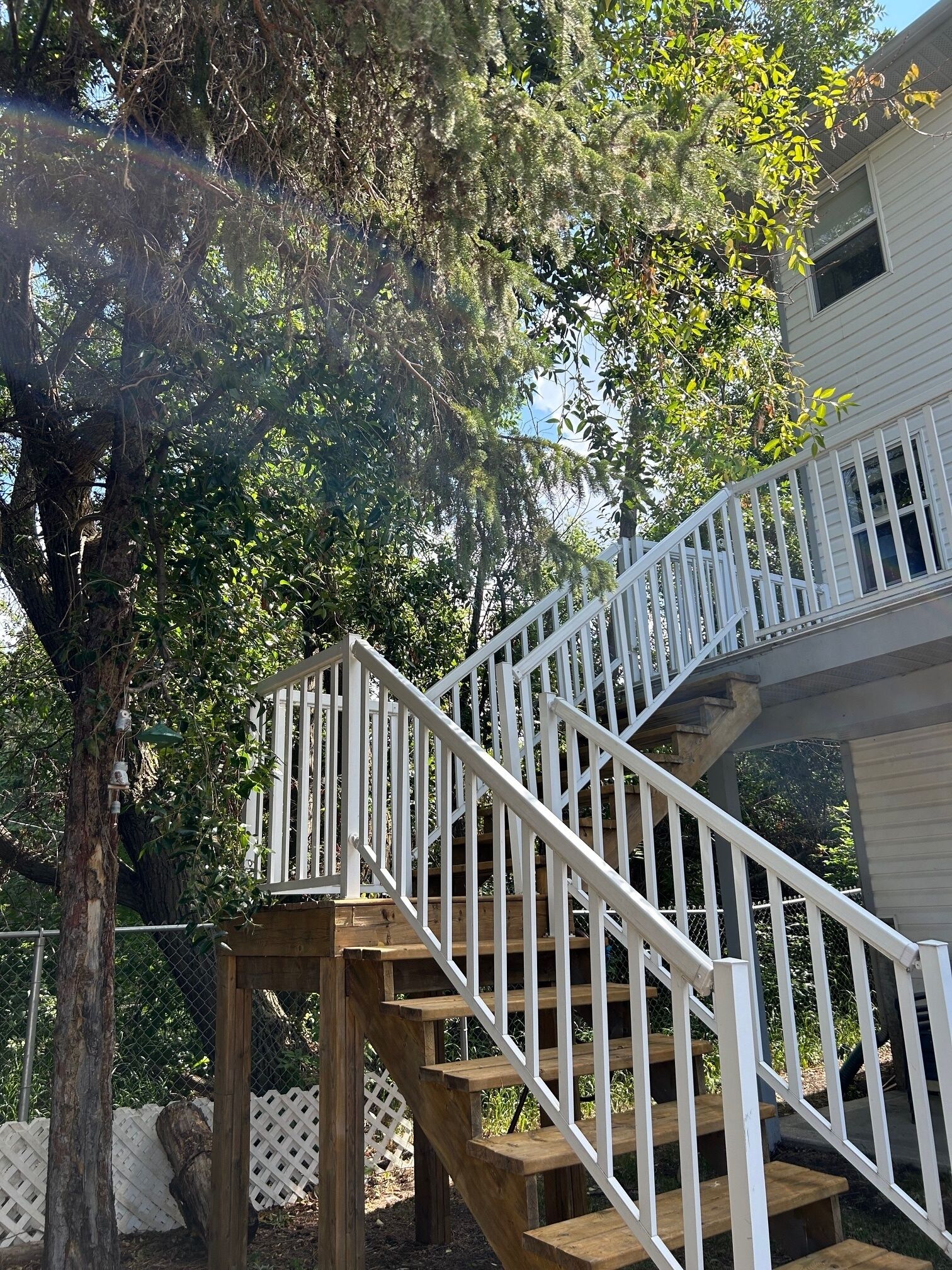 Un escalier en bois avec des rampes blanches mène à une terrasse, qui est entourée d’arbres et d’une clôture.