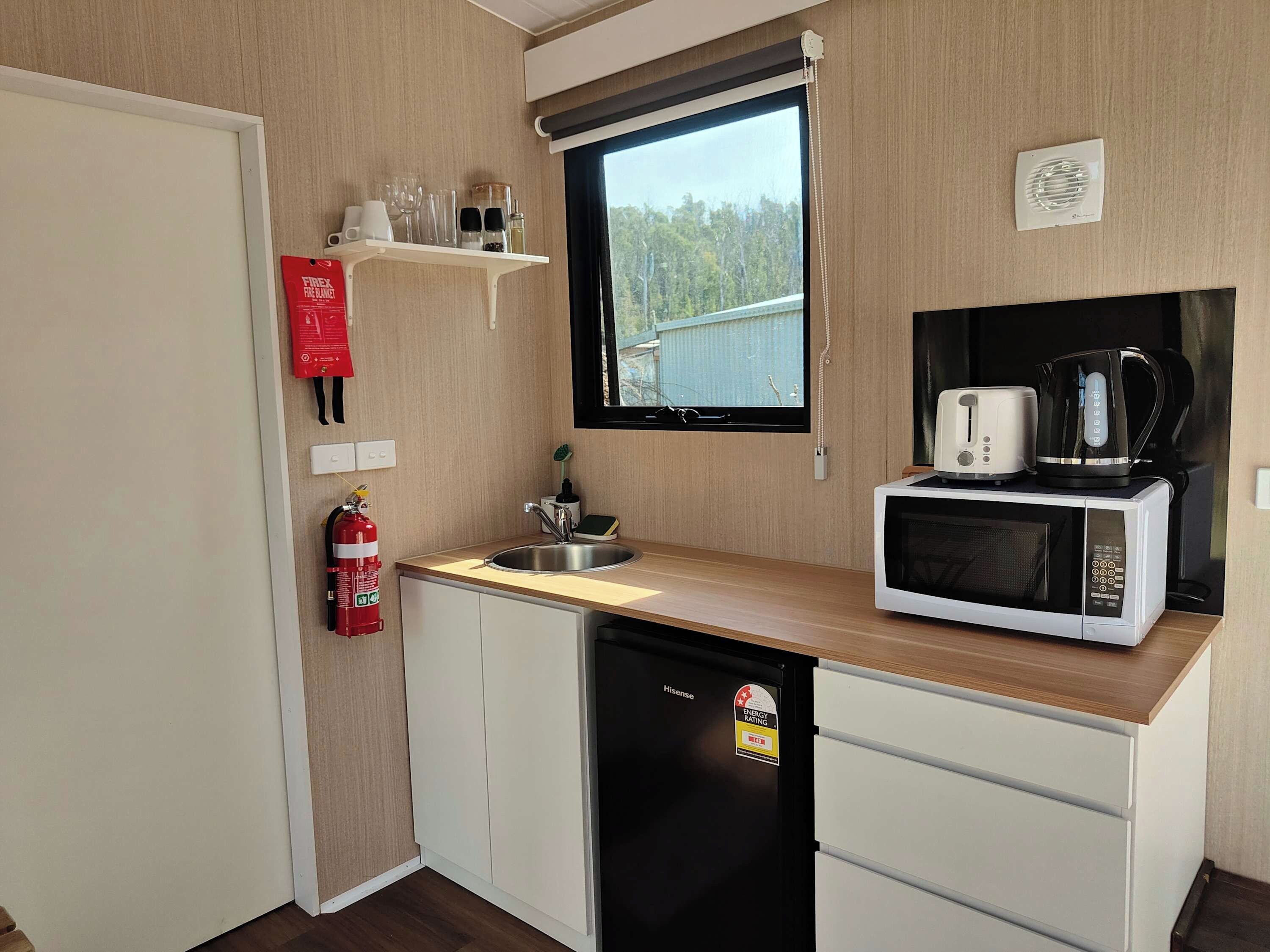 Een kleine keuken met een magnetron, broodrooster en koffiezetapparaat. Een brandblusser is aan de muur bevestigd, en een raam biedt uitzicht op de omgeving.