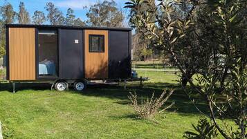 Un camper piccolo e moderno con rivestimento in legno e porta nera, parcheggiato su un prato con alberi sullo sfondo.