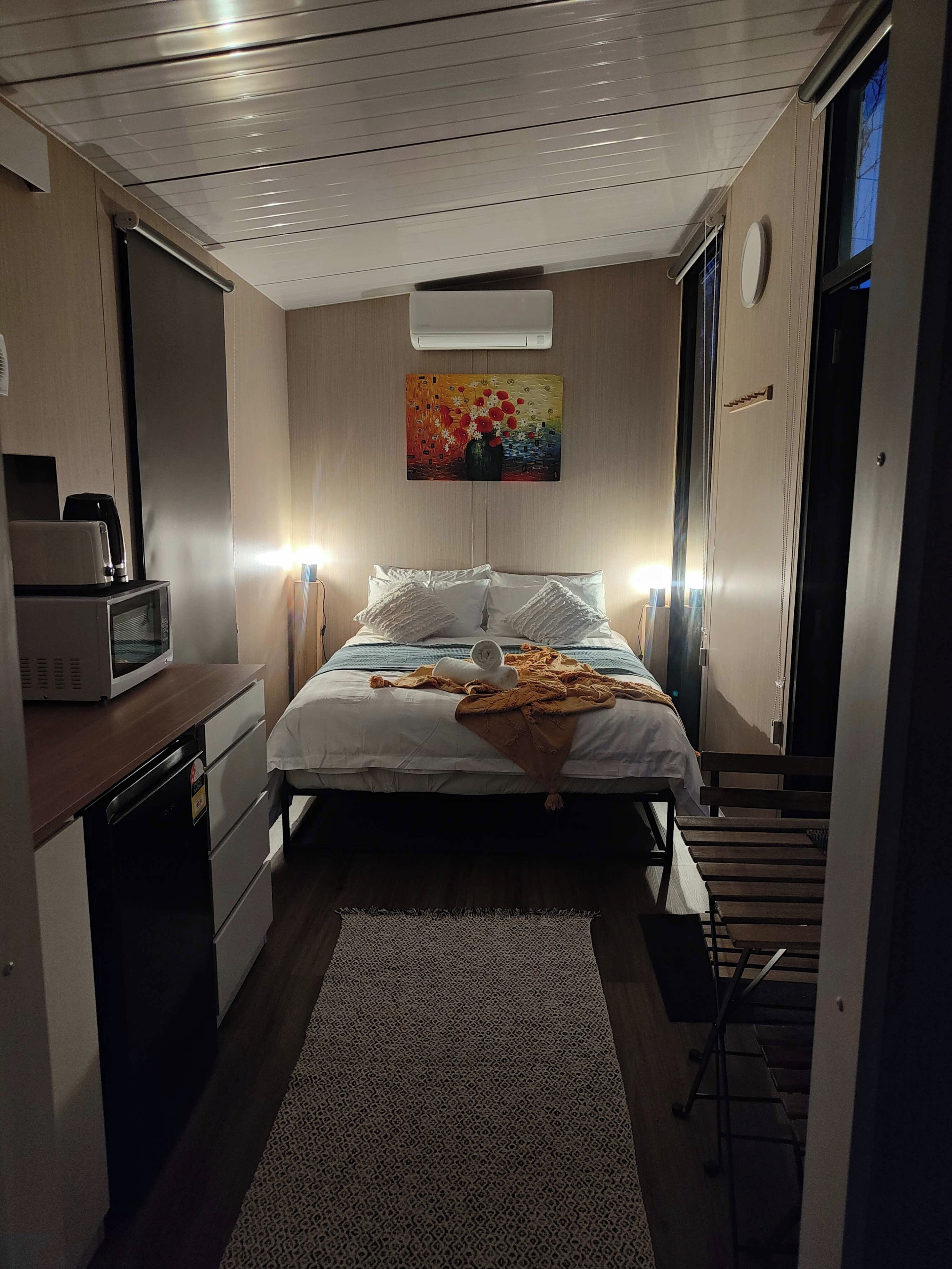 A compact hotel room with a bed, bedside lamps, a microwave, a small desk, and a painting on the wall.