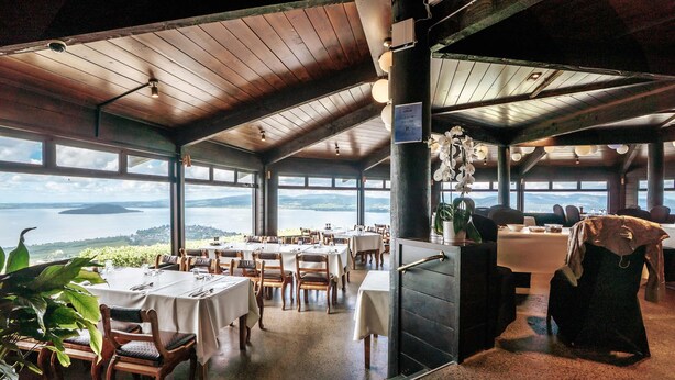 A restaurant with tables and chairs arranged for dining, large windows offering a view of the water, and a wooden ceiling. - Aorangi Peak Cabins 2 by Tiny Away (Rotorua)