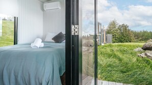 A bedroom with a bed, a window, and a view of a grassy area and trees. - Aorangi Peak Cabins 2 by Tiny Away (Rotorua)