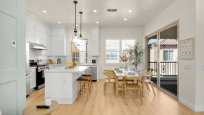A modern kitchen with white cabinetry, a central island, wooden stools, and a dining table with chairs. - Brand New Grand Luxury Designer Home in Irvine with Pool & Amenities (Irvine)