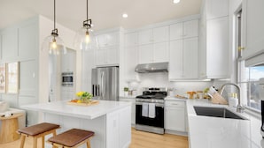 A modern kitchen with white cabinetry, stainless steel appliances, and wooden stools. - Brand New Grand Luxury Designer Home in Irvine with Pool & Amenities (Irvine)