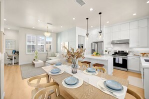 Una cocina moderna con armarios blancos, un comedor con mesa de madera dispuesta para cuatro personas, y una sala de estar con sofá y una mesita de centro.