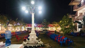 An outdoor seating area with rows of plastic chairs and tables, a central decorative pillar, and a water dispenser. - The Narayani Resort (Gaindakot)