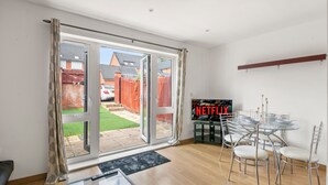 A modern living room with a glass door leading to a garden, a dining area with a glass table and chairs, and a Netflix logo on the wall.