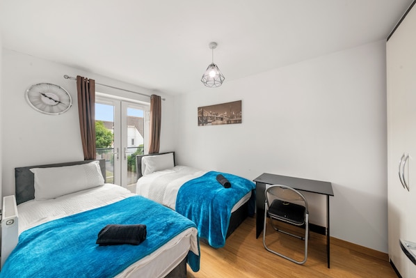 Ett sovrum med två sängar, ett skrivbord, en stol, en väckarklocka och en tavla som föreställer en stadsbild.