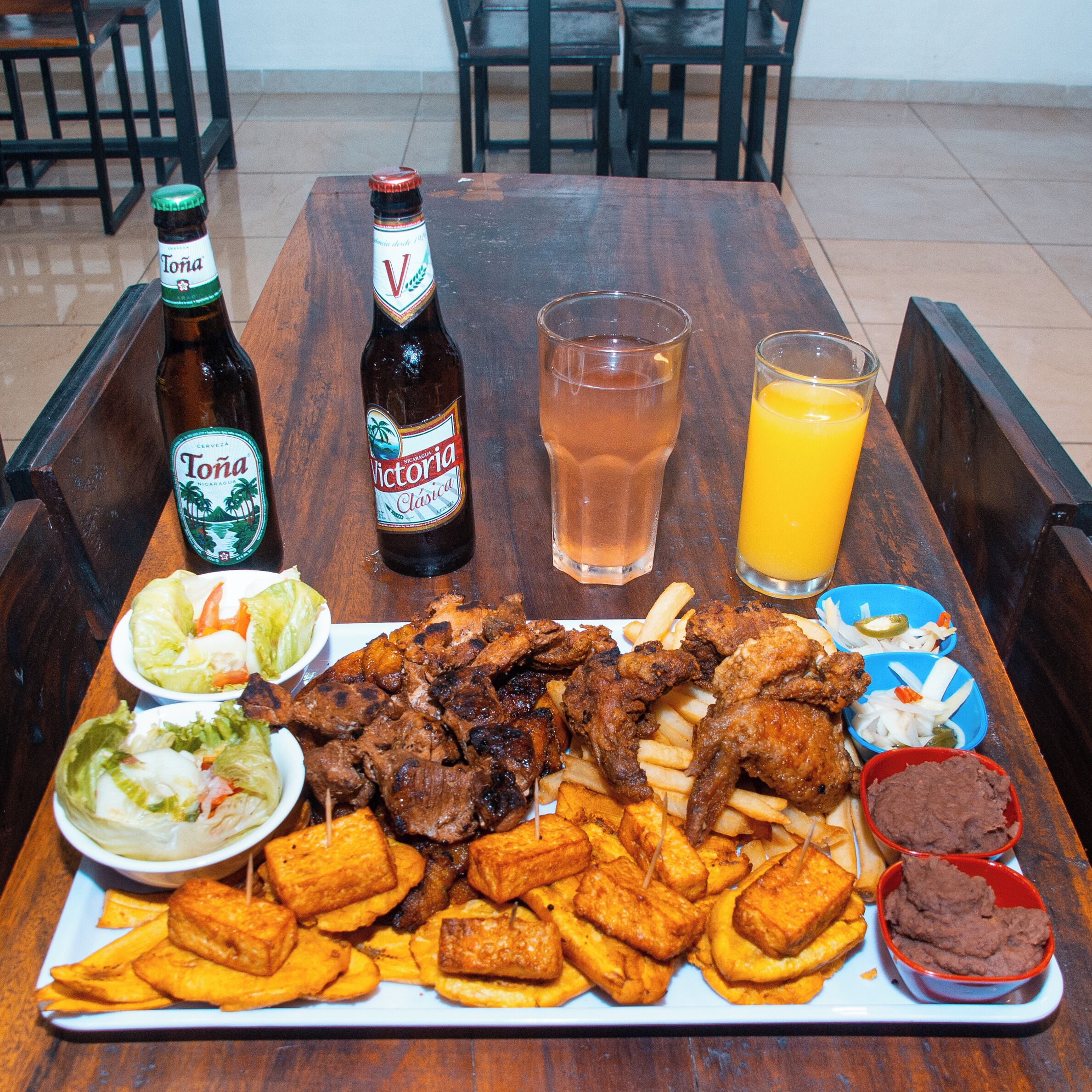 Una mesa de madera con comida que incluye carne a la parrilla, papas fritas y acompañamientos, además de dos botellas de cerveza y un vaso de jugo.