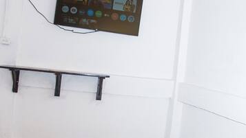 Quarto com cama, televisão fixada na parede exibindo diversos canais e prateleira de madeira.