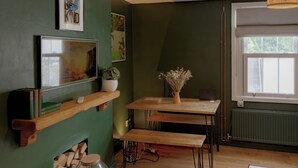 Una habitación con pared verde, una mesa de madera con bancos, una chimenea y una ventana con vistas a la vegetación.