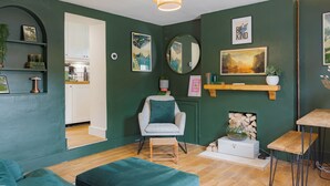 Una habitación con una pared verde, suelo de madera, un sofá verde, una silla, una mesita y un espejo.