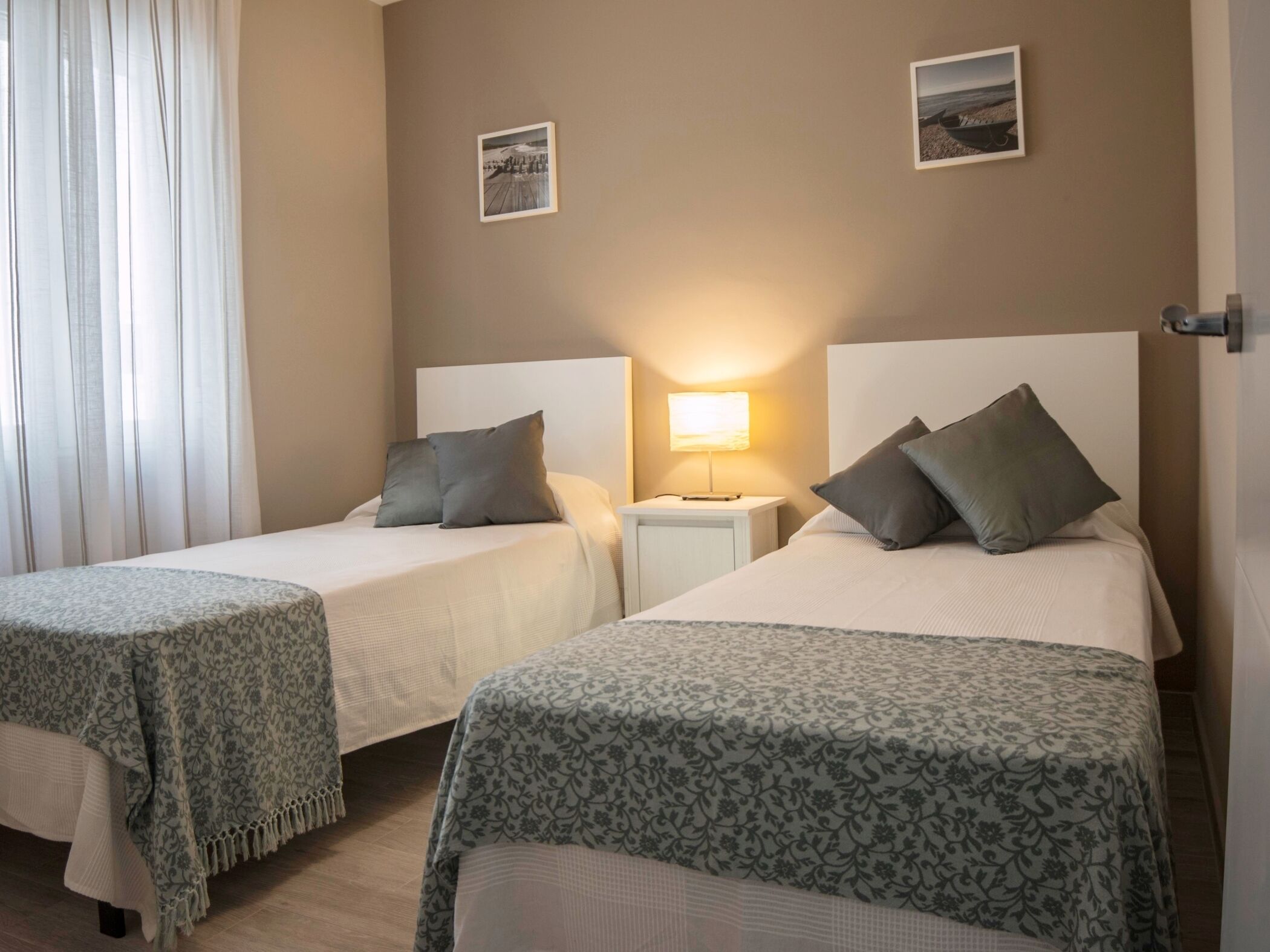 A hotel room with two beds, each with a headboard, a bedside table with a lamp, and a window with curtains.