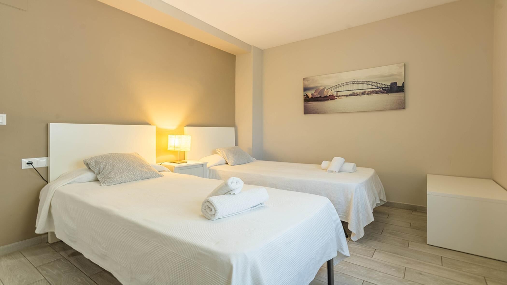 Une chambre d’hôtel avec deux lits, une table de chevet avec une lampe, un tableau représentant un pont et une petite table avec des serviettes.