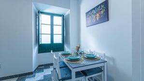 Un coin repas avec une table dressée pour quatre personnes, une fenêtre avec un volet vert et un tableau accroché au mur.