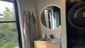 A modern bathroom with a built-in washing machine, a wooden vanity with a mirror, and a view of greenery outside.