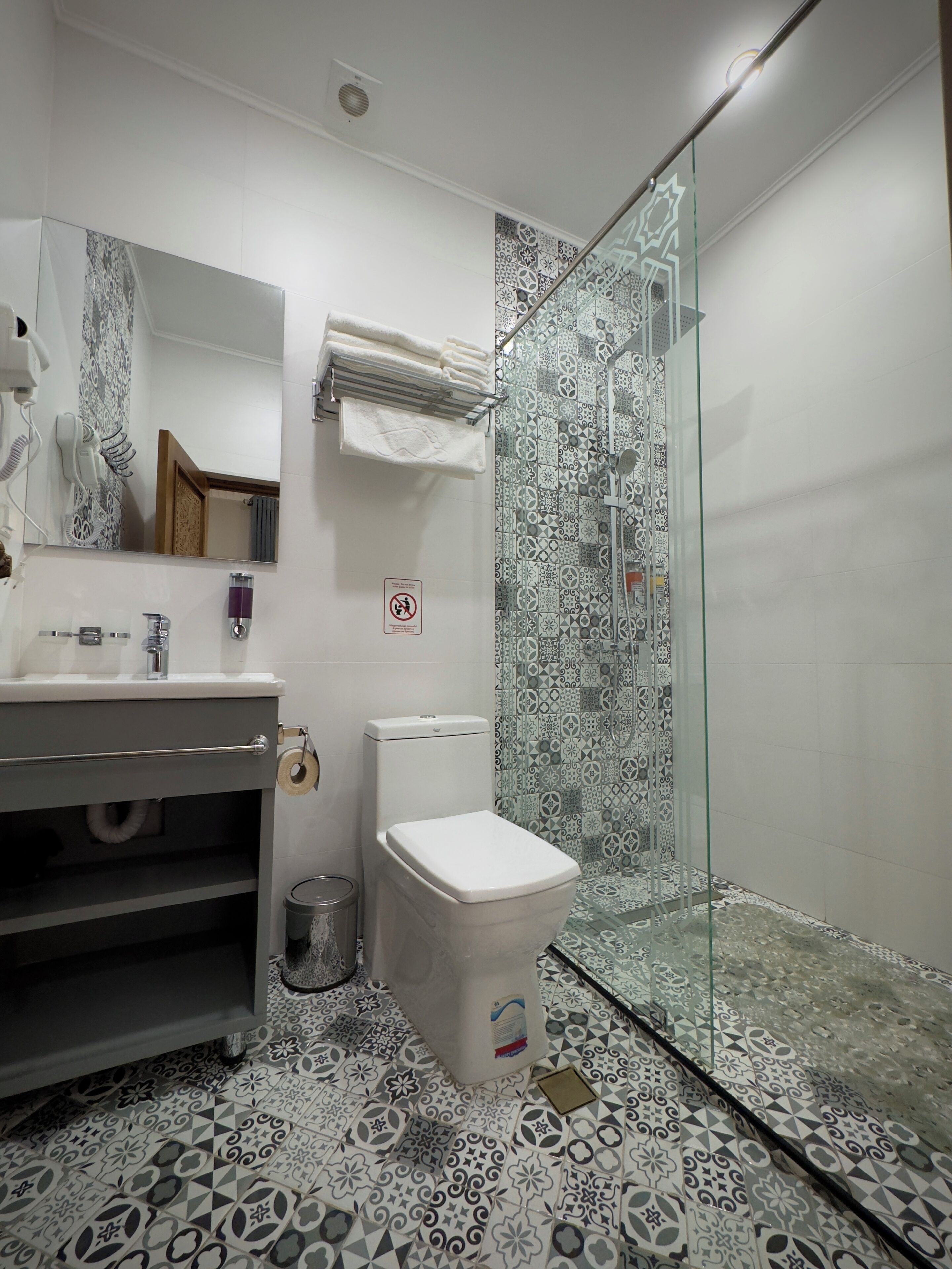 A modern bathroom with a glass shower enclosure, a white toilet, a sink with a mirror, and patterned floor tiles.