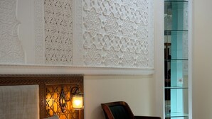 Habitación de hotel con cama, una silla, una pared con intrincados grabados, una mesita de noche con lámpara y un aparador de vidrio.