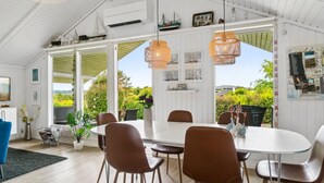 A dining area with a white table and brown chairs, large windows with a view of greenery, and a ceiling with exposed beams and hanging lights.