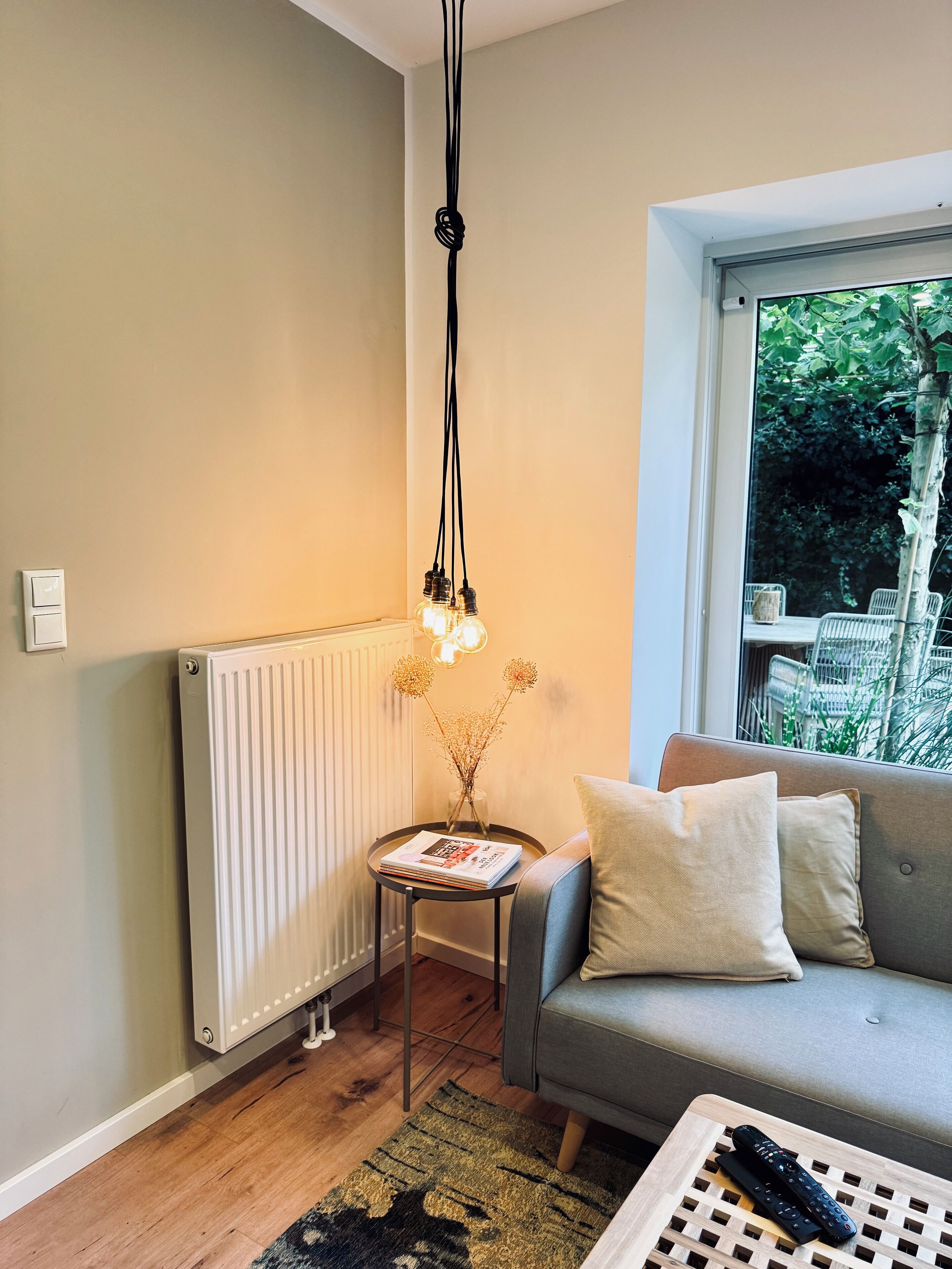 A cozy living room with a grey sofa, a small side table with a magazine, a radiator, and a hanging light fixture.