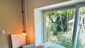 A bedroom with a bed, a bedside table with a tray of fruit, a radiator, and a window with a view of greenery.