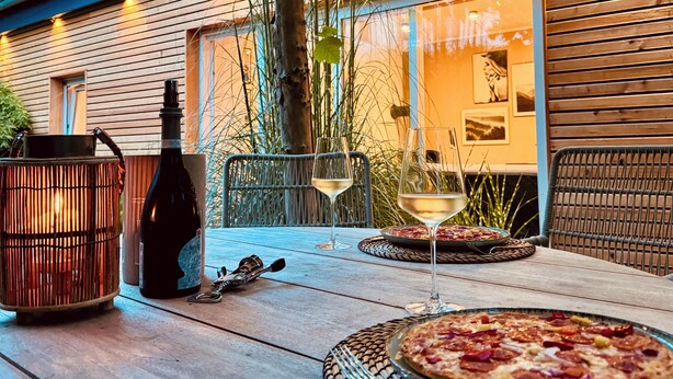 An outdoor dining area with a table set for two, featuring a pizza and wine glasses.