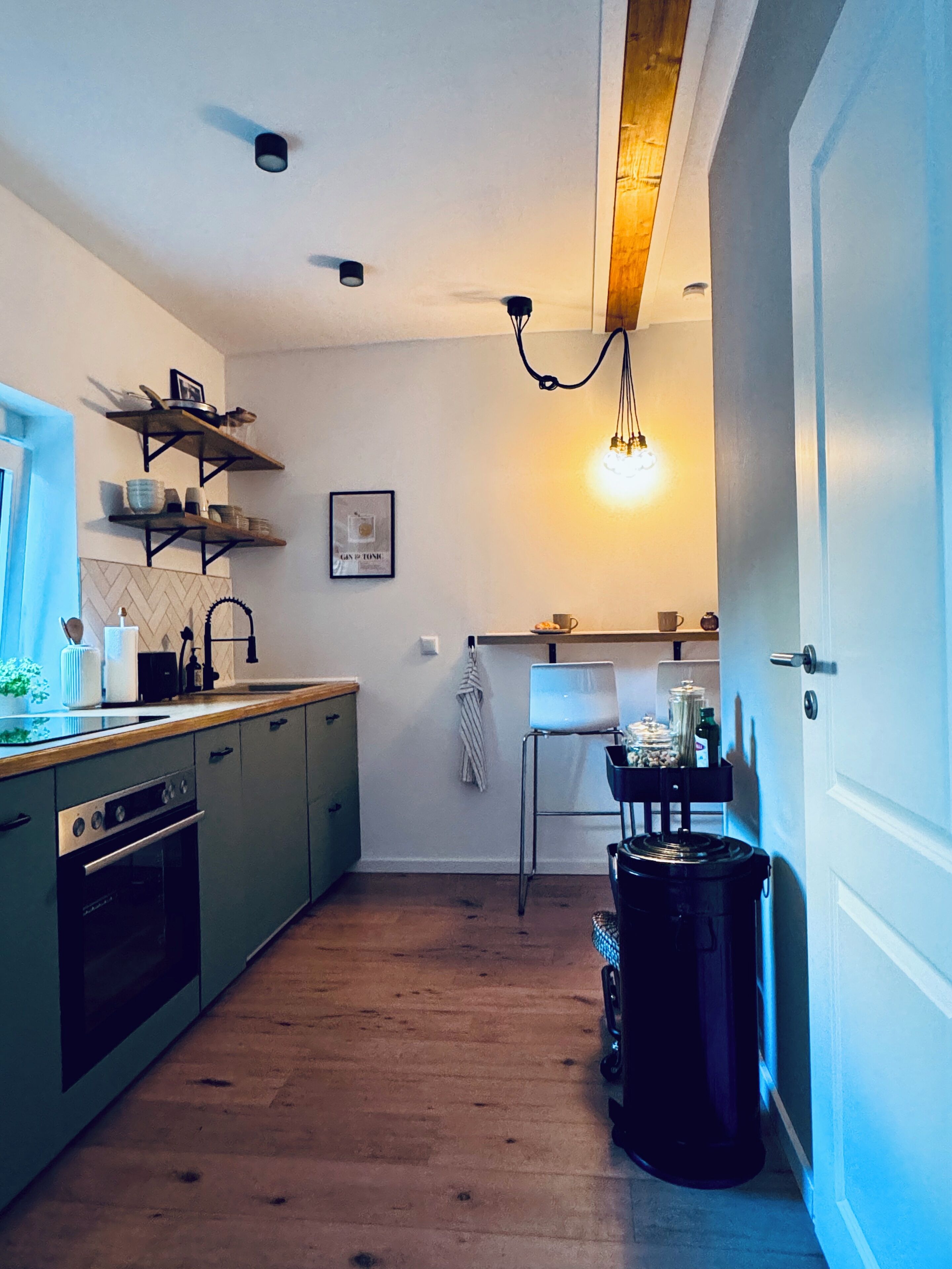 Una cucina compatta con forno a incasso, lavello e piano di lavoro in legno. Si vede una sedia e un cestino per i rifiuti vicino alla porta.