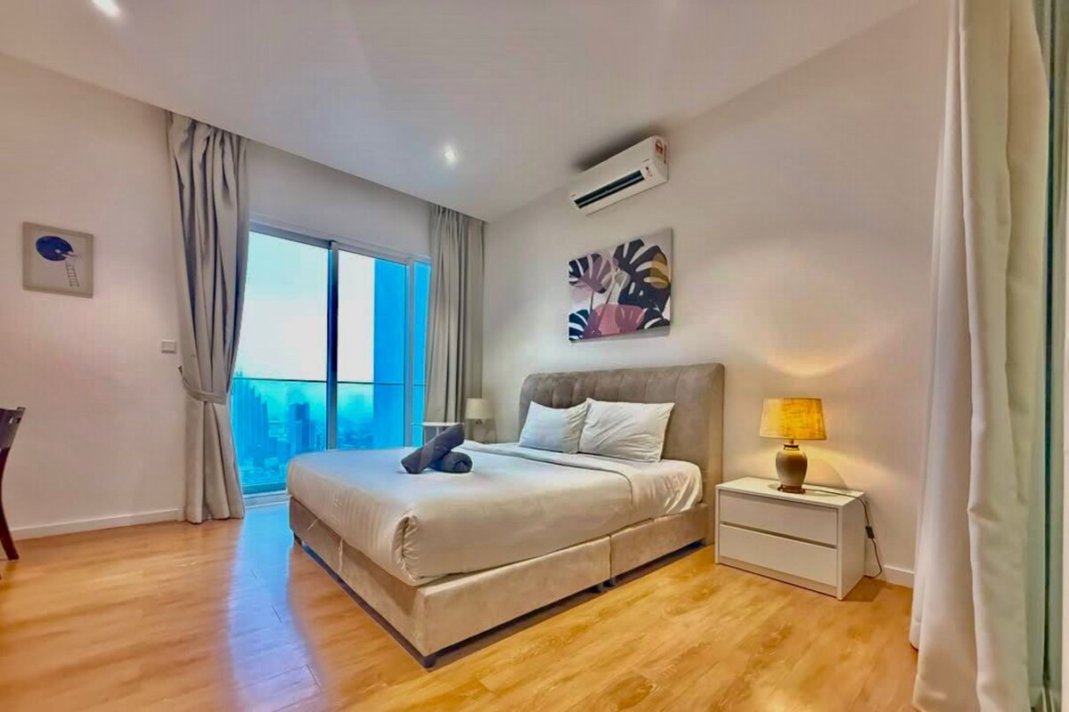 A modern bedroom with a large bed, a bedside table with a lamp, and a view of the city through a large window.