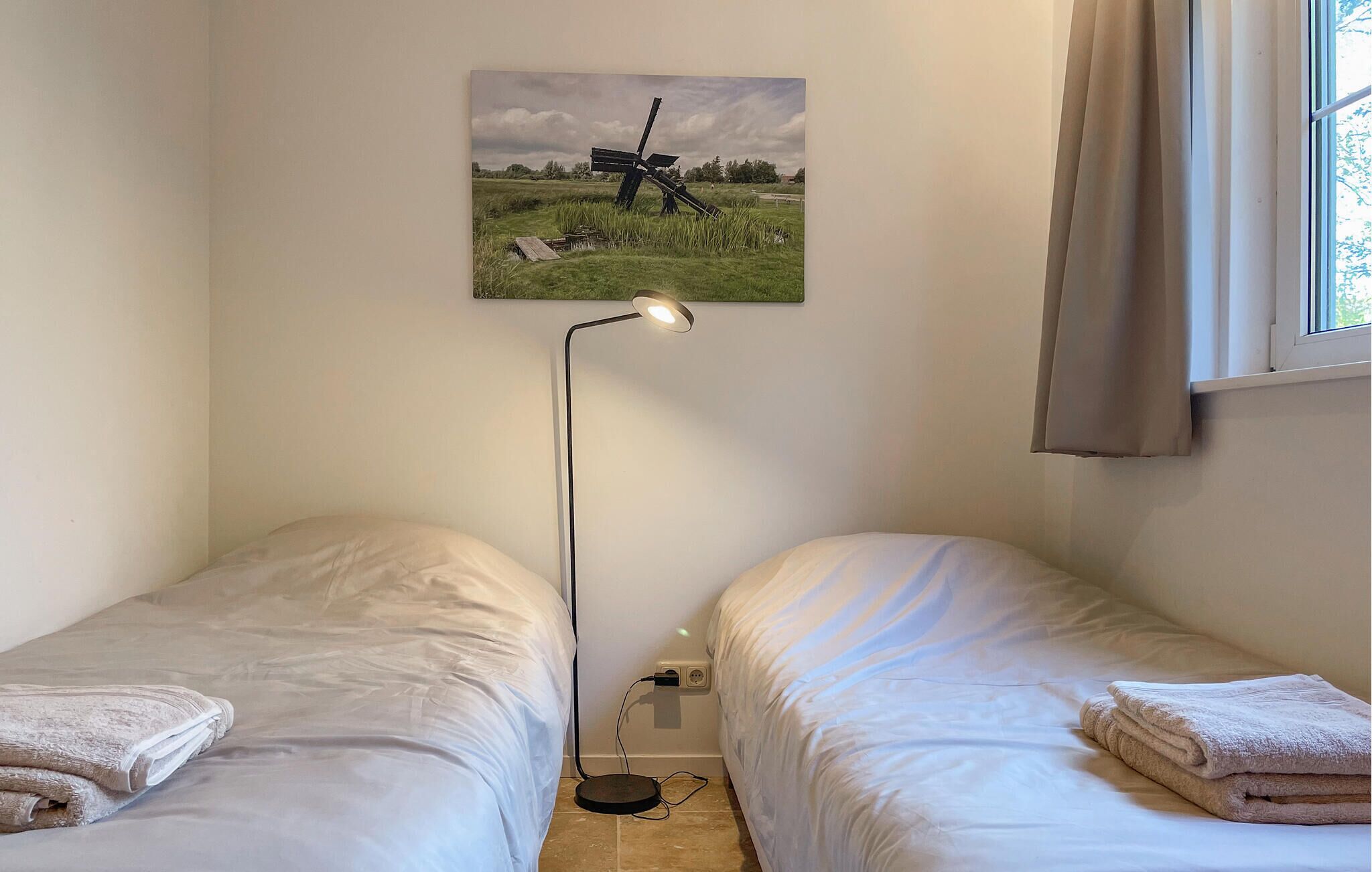 A bedroom with two single beds, a floor lamp, a painting of a windmill, and a window with curtains.