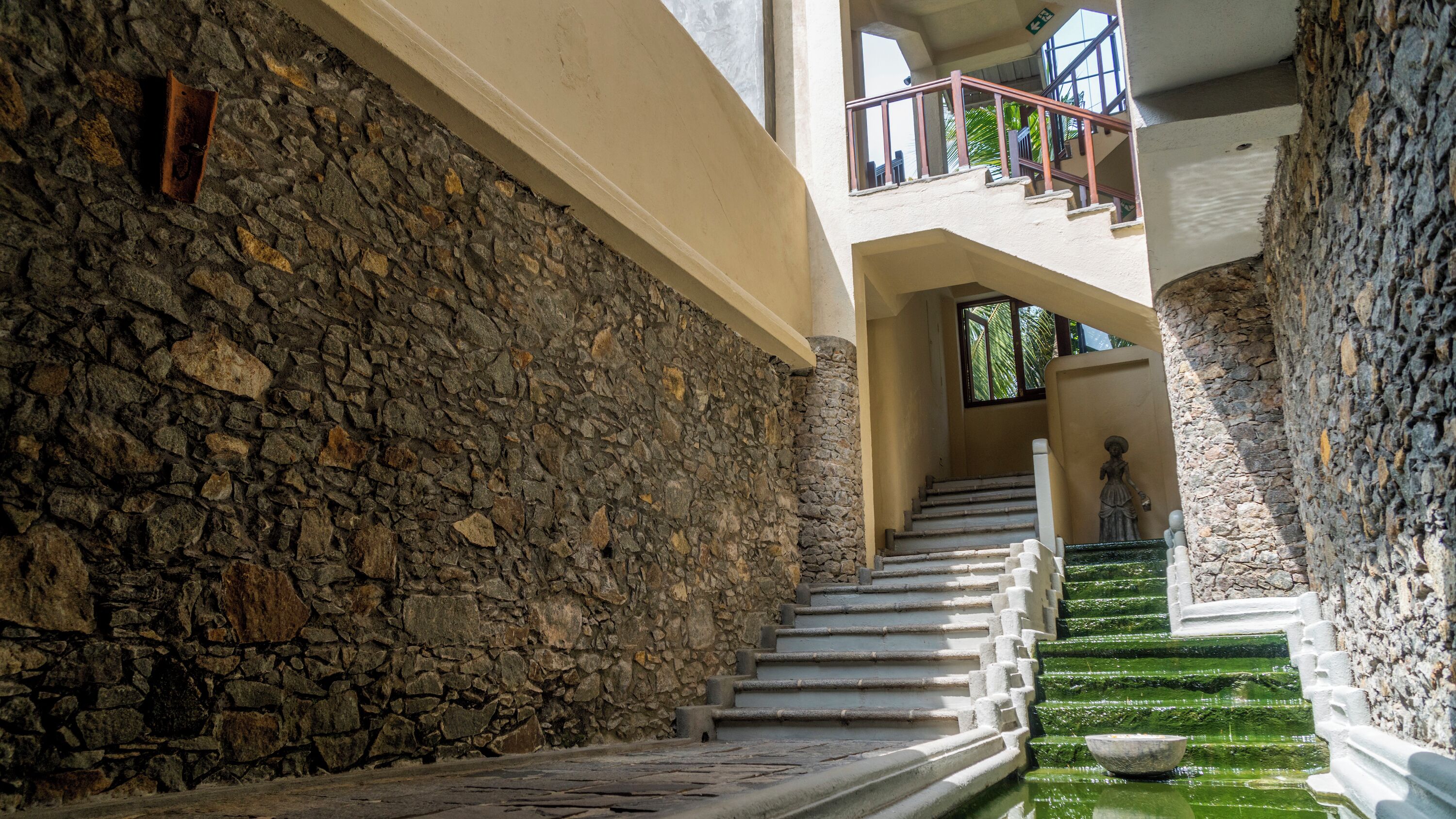A stone staircase leading to a balcony with a statue, surrounded by stone walls and a water feature.