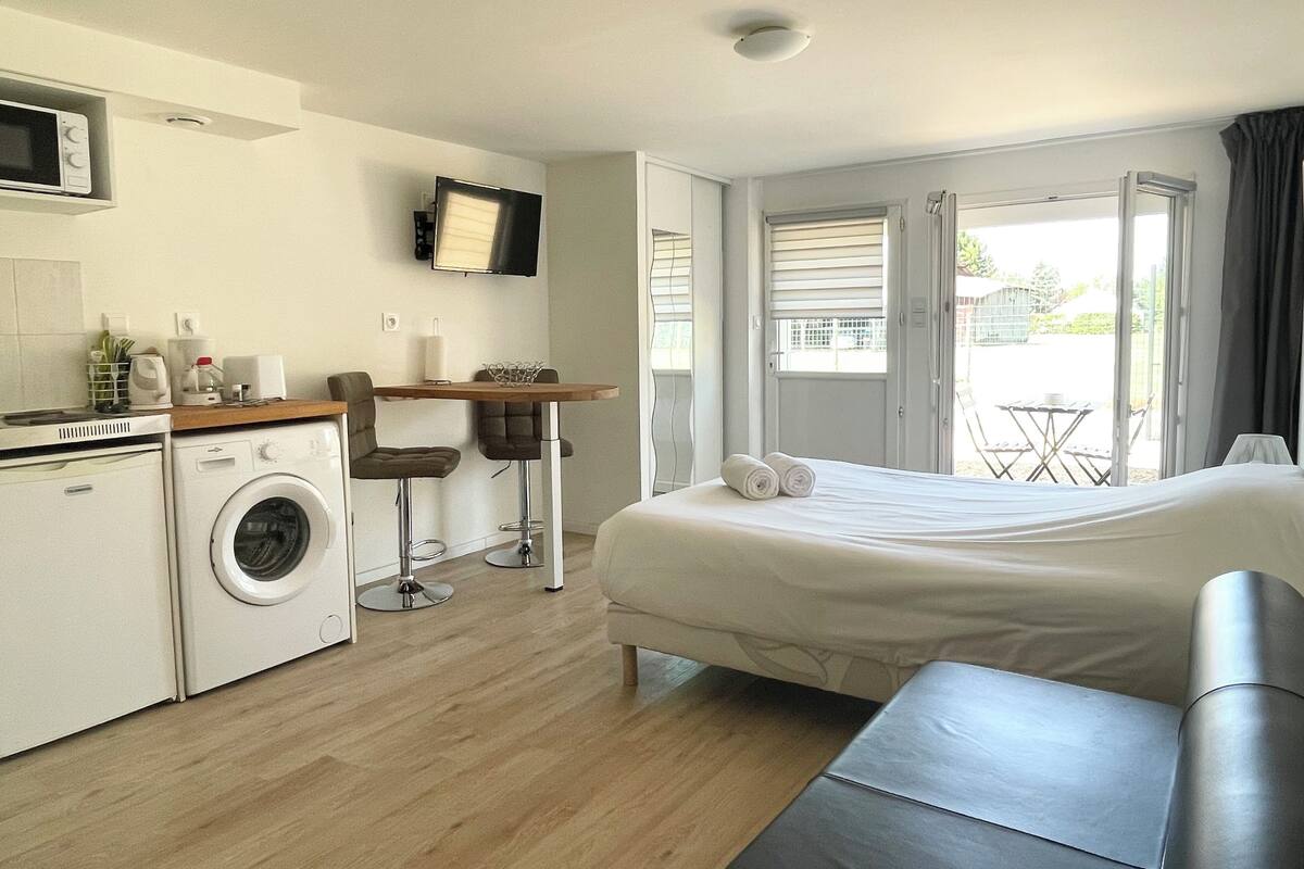 Habitación de hotel compacta con cama, kitchenette pequeña, lavadora, televisor y balcón con mesa y sillas.
